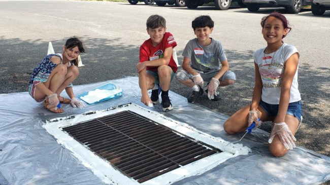 four students sit around a drain on the street with cars in the background. The students are wearing gloves and holding paint brushes. Around the drain is fresh white paint.