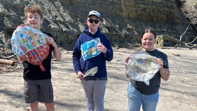 Three smiling children stand on a beach holding deflated mylar balloons.