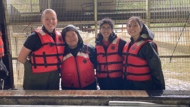 Four people in life jackets and waders, smiling and posing together. They're standing in in knee high water in a fish hatchery.