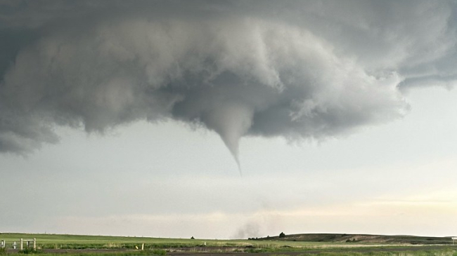 Dark clouds swirl together at the top of the frame and a tornado cone begins to form below the clouds above a cornfield. Flat farmland extends as far as the eye can see; on the horizon, the sun is showing through lighter clouds.