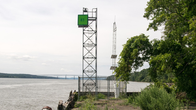 A tower with sensors sits on a shoreline with a bridge and trees in the background