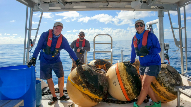 Three people wearing life preservers standing with large spherical yellow buoys on a boat in the ocean
