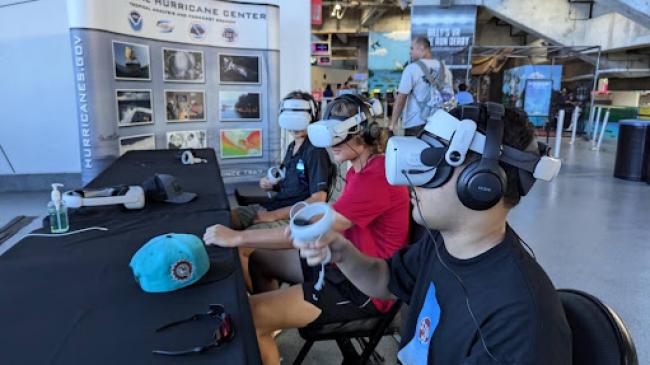 People at a public sporting event try out the Weather the Storm virtual reality storm surge simulator.