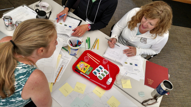 Educators are engaged in conversation while seated at a table with art supplies, pens, post-its, and notebooks. 