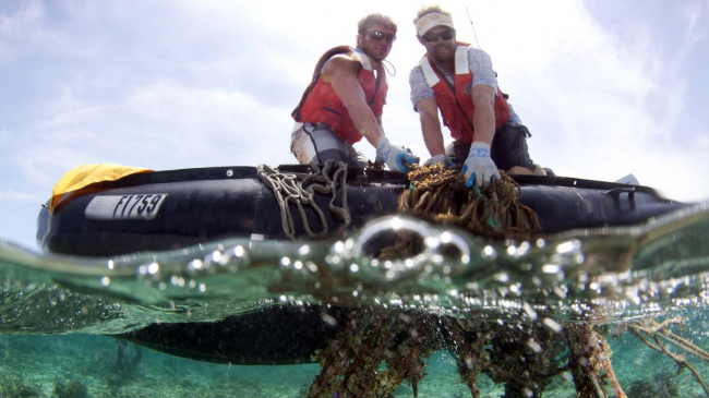 Two people wearing orange life vests on a floating raft pull up large rope from the ocean.