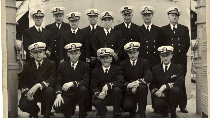 A group of NOAA Corps officers posing for a photo in the wardroom of the Coast and Geodetic Survey Ship Explorer. Eight men are standing and five more men are kneeling in front of them.