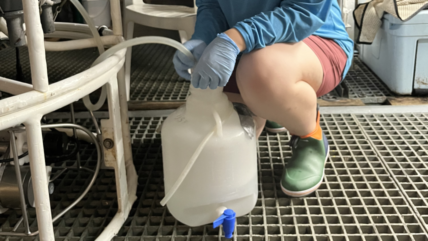Anna smiles and kneels on the grated vessel floor. In a rubber gloved hand, Anna holds a tube coming from a large sampling device into the opening of a plastic jug with a spigot at the bottom.