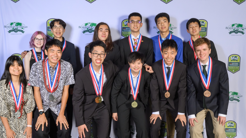 Twelve students dressed in formal attire pose for a group photo in front of a backdrop that has the 40th Science Olympiad National Tournament logo on it.
