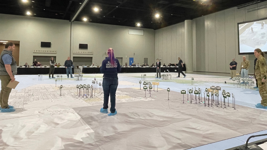 A group of adults standing in a large room stand around small flags on the ground representing response resources on a 65x65ft map of Alaska.