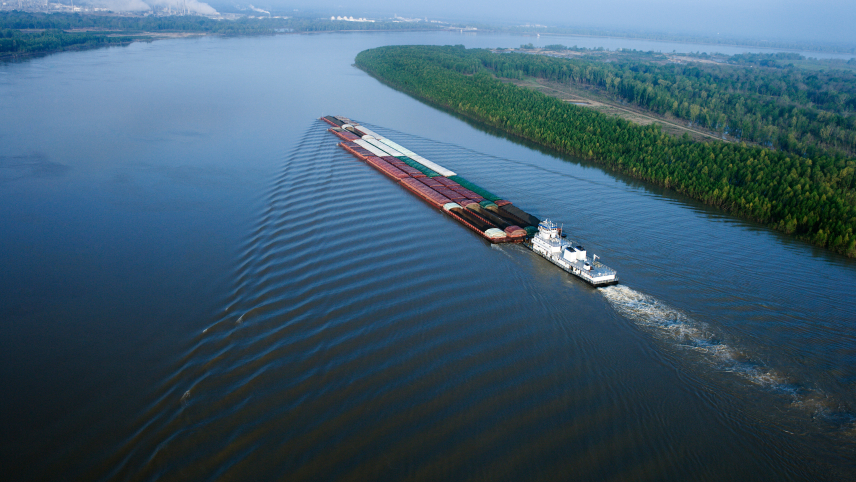 A long ship carrying cargo goes down a wide river surrounded by green landscape on either side