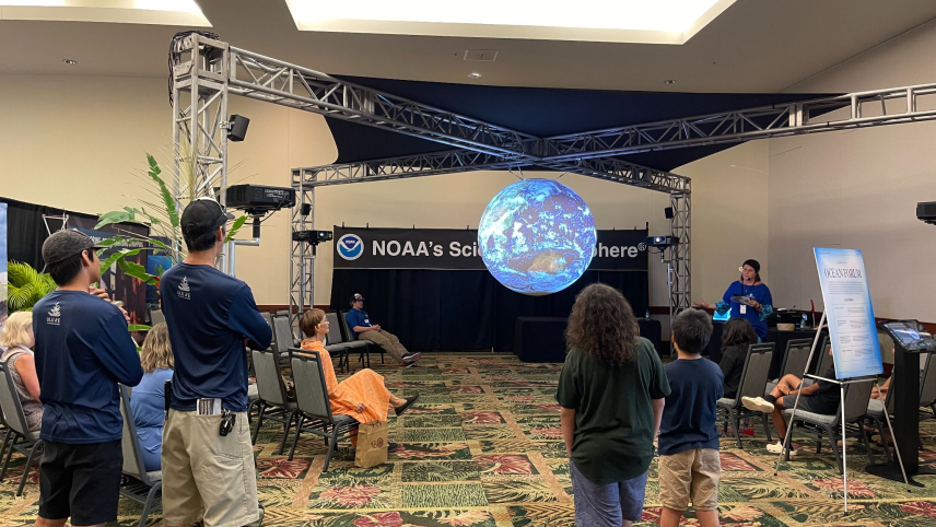  A group of people are standing or sitting in a room displaying a big three dimensional sphere displaying the Earth. 