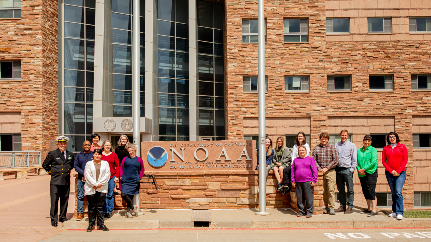 A group of adults stand outside a brown building. Seven adults stand left and eight adults stand to the right of a sign with the NOAA logo. The top line of the sign reads “U.S. Department of Commerce”, the middle reads “NOAA”, and the bottom reads “National Oceanic and Atmospheric Administration David Skaggs Research Center.”  