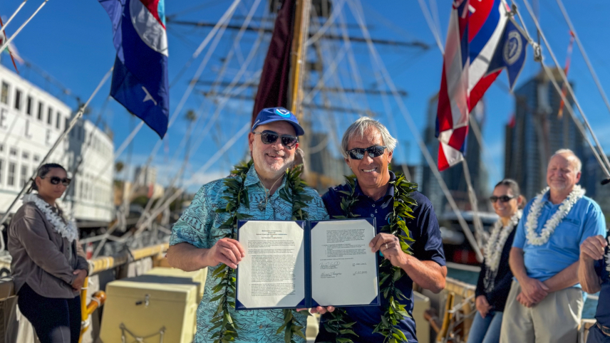 Two people with green leis wrapped around their necks stand smiling and posing on a canoe outside holding up a folder with papers. A few other people are standing on either side of them. Two flags hang from either side of the canoe as well.