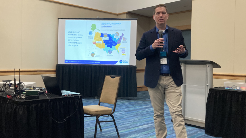 a man speaking inside a room between two  tables with black table covers. There is a chair and a podium behind him as well as a projection screen displaying a colorful map.