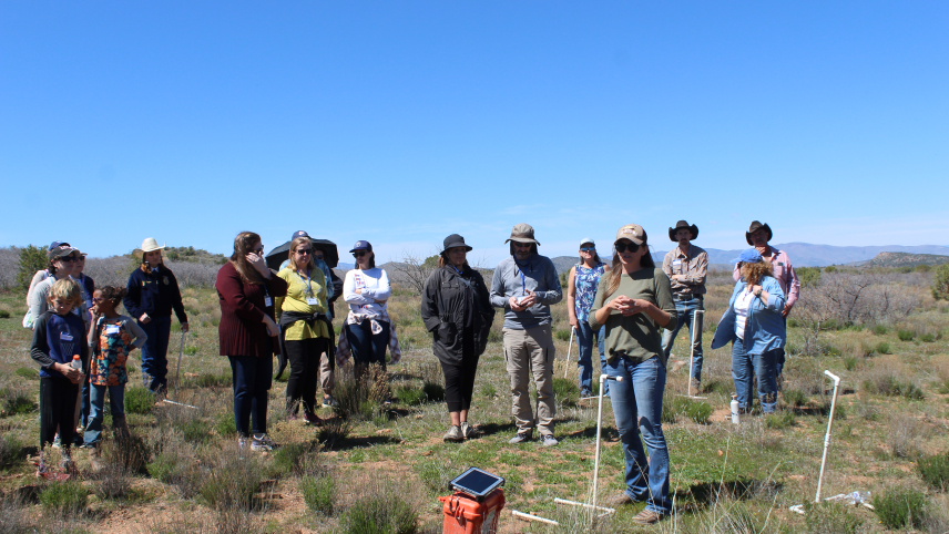 A group of people gathered outdoors to observe a speaker using agricultural measuring tools. 