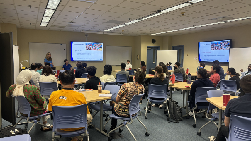 people in a conference room sitting at tables and looking in the direction of screens and presenters.