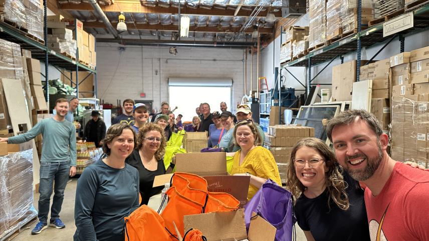 Group of people posing by a table and smiling, in a warehouse with tall shelves and boxes around them.