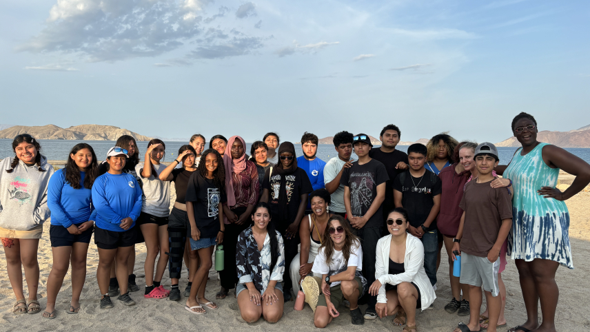 Twenty-seven males and females stand on beach sand with a body of water and islands behind them. 