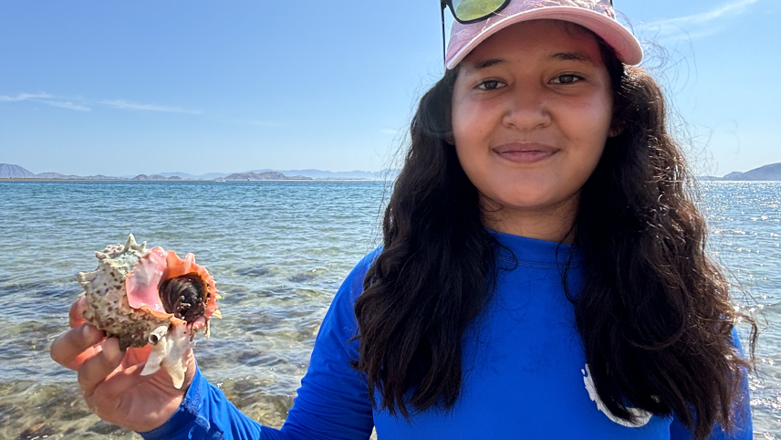 A teenager poses outside next to the ocean in a blue long-sleeve shirt holding a white, brown, and pink conch shell containing a brown sea snail emerging from the shell’s opening. 