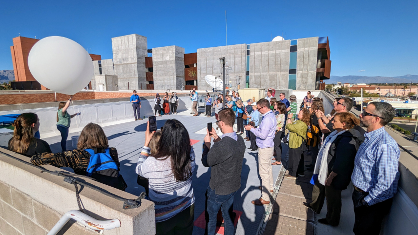 A group of people gathered on the rooftop observing and taking pictures of a staff with a large white balloon about to be released in the sky. 