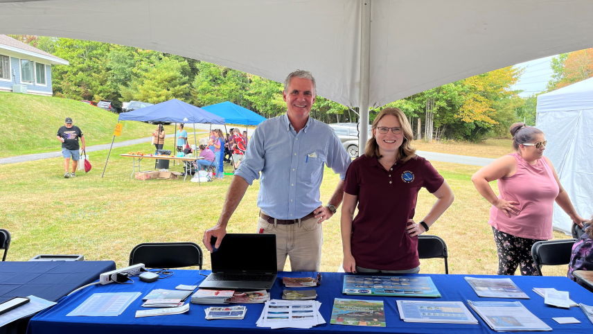 Two adults at an outdoor event, standing behind an informational table under a tent.