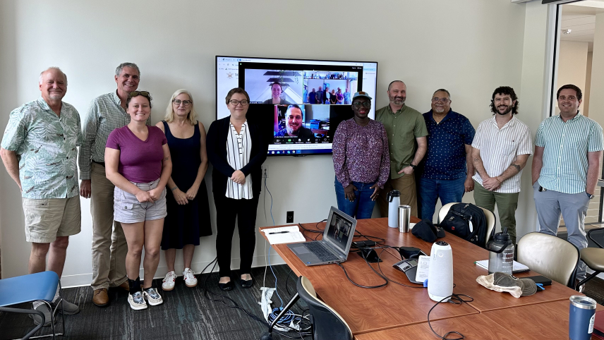 Group of adults posing in a conference room by a screen with faces of people who joined virtually on it. 