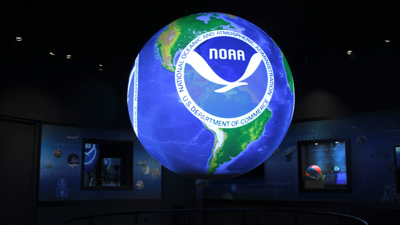 A Science on a Sphere in a dark room. The Sphere has a map of the world with a large NOAA logo on top.