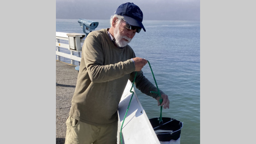 Clive is standing on a pier, wearing brown pants, a brown sweater, and a blue baseball cap, hoisting a net from over the fence. He is leaning against a white fence with the ocean just beyond him.