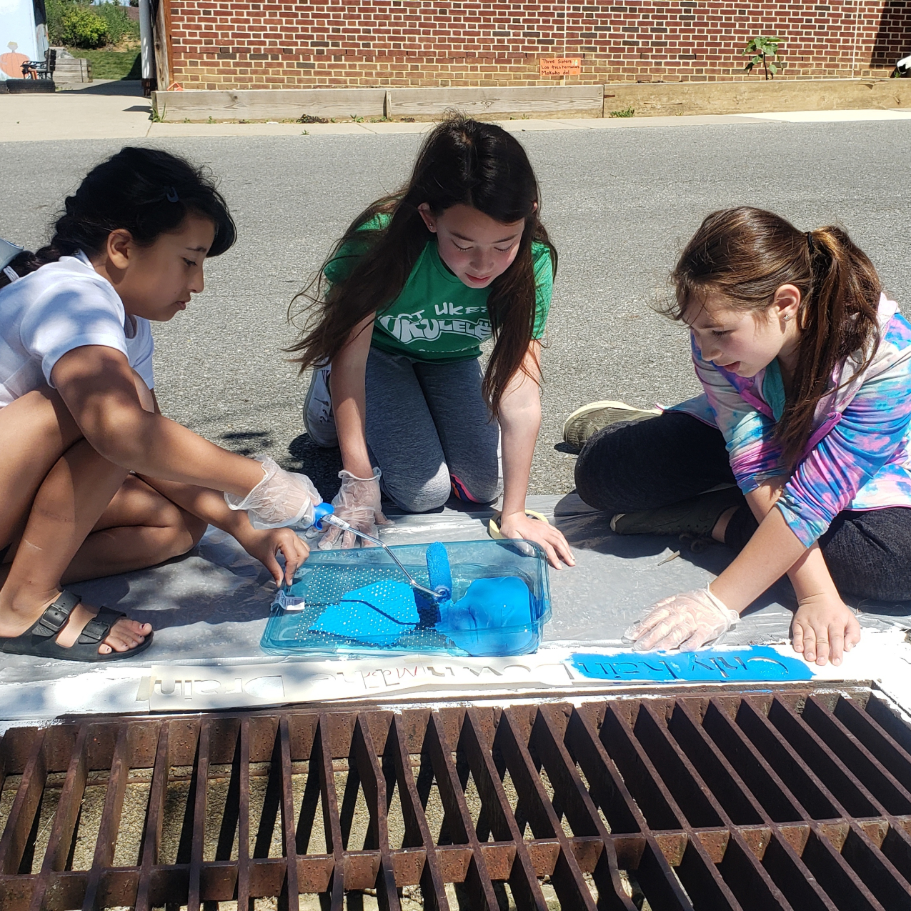 Three kids sit next to a storm drain along a road with a brick building in the background. The storm drain has white paint bordering the drain. The kids have a stencil on top of the white paint, and are painting blue on top of the stencil to create messaging around the storm drain.