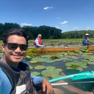 A selfie-style photo taken by Keanu. He smiles in the foreground in his kayak. Two people are in a canoe in the background and smile for the camera. Everyone is wearing protective life vests and sun protection as they float among the lotus and lily pads on a calm sunny day.
