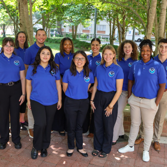 Twelve people pose and smile for the camera outside. They are wearing blue polo shirts that have a NOAA logo in the upper right. 