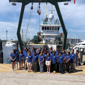 A group of people in matching blue shirts with CCME logos pose in front of a large, docked vessel.