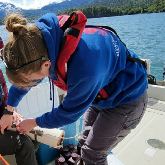 Seraina and Nicole are aboard a small research vessel on what looks to be calm waters. Seraina is manipulating a cylindrical instrument that Nicole holds in her lap. They are dressed for cold weather fieldwork and wear personal floatation devices.