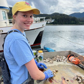Kate smiles, standing at a muddy table on a dock. She uses gloved hands to grab oysters and her full-length waders are covered in mud from the task.