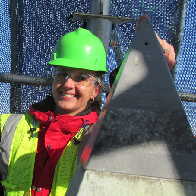National Geodetic Survey Director, Juliana Blackwell, touches the top of the Washington Monument with her forefinger. She stands on scaffolding surrounding the monument, wearing a hard hat and a safety vest.