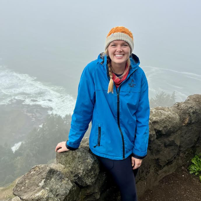 A woman in a raincoat and hat stands on a cliff near a foggy coastline.