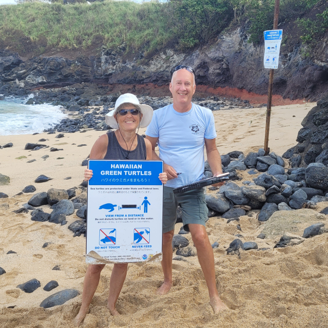 Jerry and Esther stand together on a beach in Hawaii. Jerry is holding a clipboard and Esther is holding a sign with information about sea turtle nesting.
