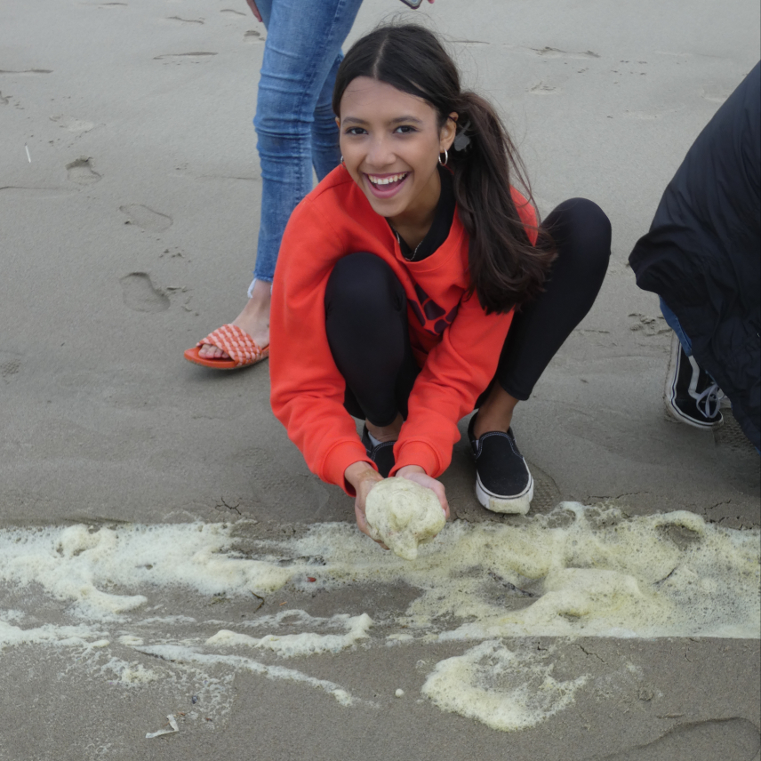 A student crouching down in front of sea foam holding a handful of foam and smiling at the camera.
