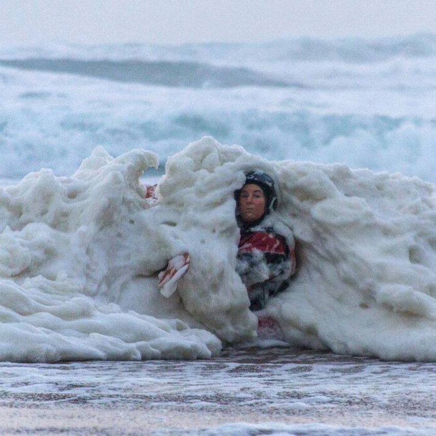 A wave full of thick sea foam crashes over a kayaker. Only the kayaker's face, shoulder, and half of a paddle is visible through the foam.